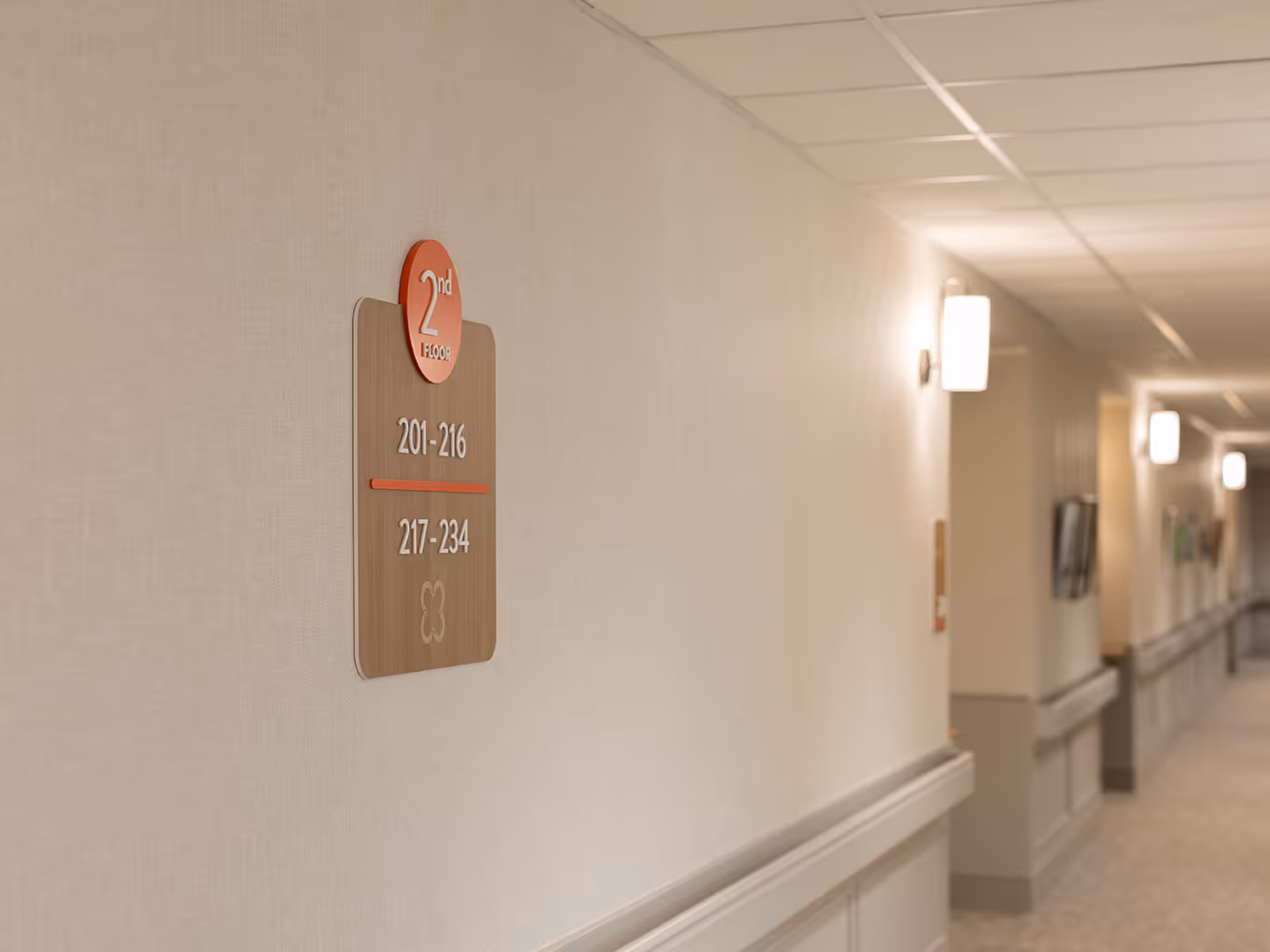 Hallway view at United Active Living Garrison Green showing the second-floor wing identifier sign with a coral floor badge and suite range markers, set against a warmly lit corridor.