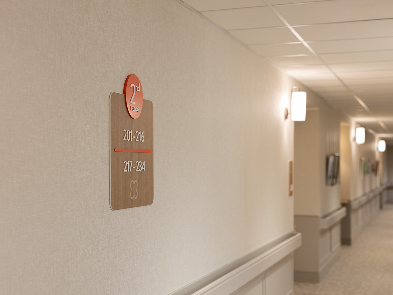 Hallway view at United Active Living Garrison Green showing the second-floor wing identifier sign with a coral floor badge and suite range markers, set against a warmly lit corridor.
