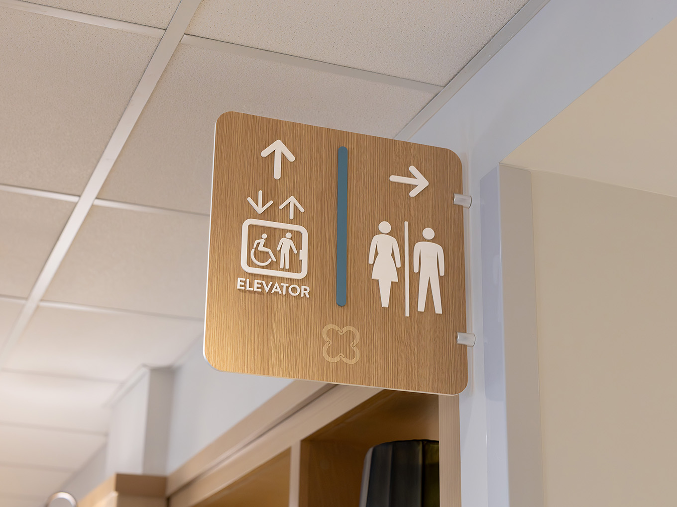 Wall-mounted elevator and washroom directional sign in a hallway at United Active Living Garrison Green, featuring custom icons and a coordinated wood-look finish.