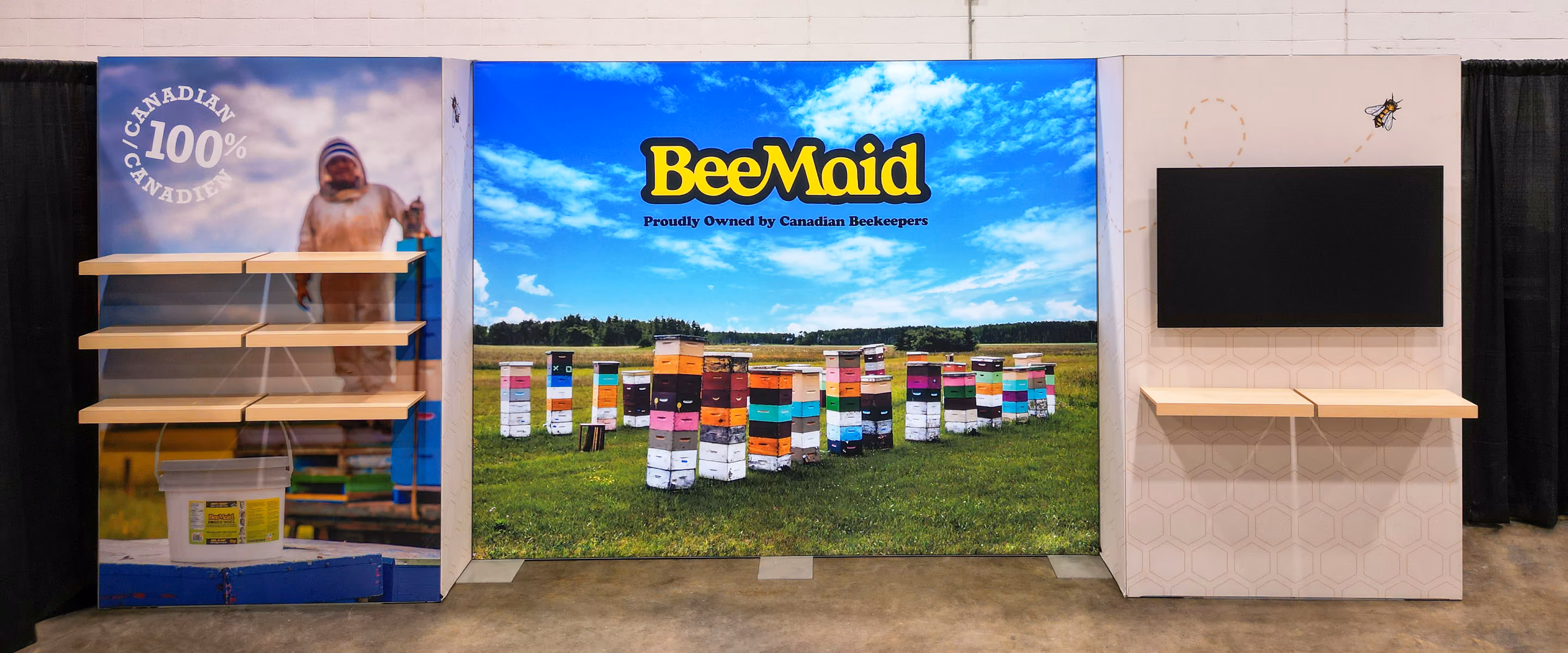BeeMaid Honey 10x20 trade show display featuring large-format backlit graphic backdrop, beehive photo mural, beekeeper panel with product shelving, and monitor wall.