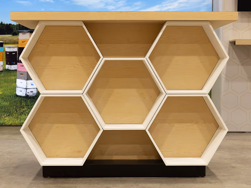 Front-facing view of custom white and maple hexagonal honeycomb product display counter — fabricated trade show furniture for BeeMaid Honey brand activation.