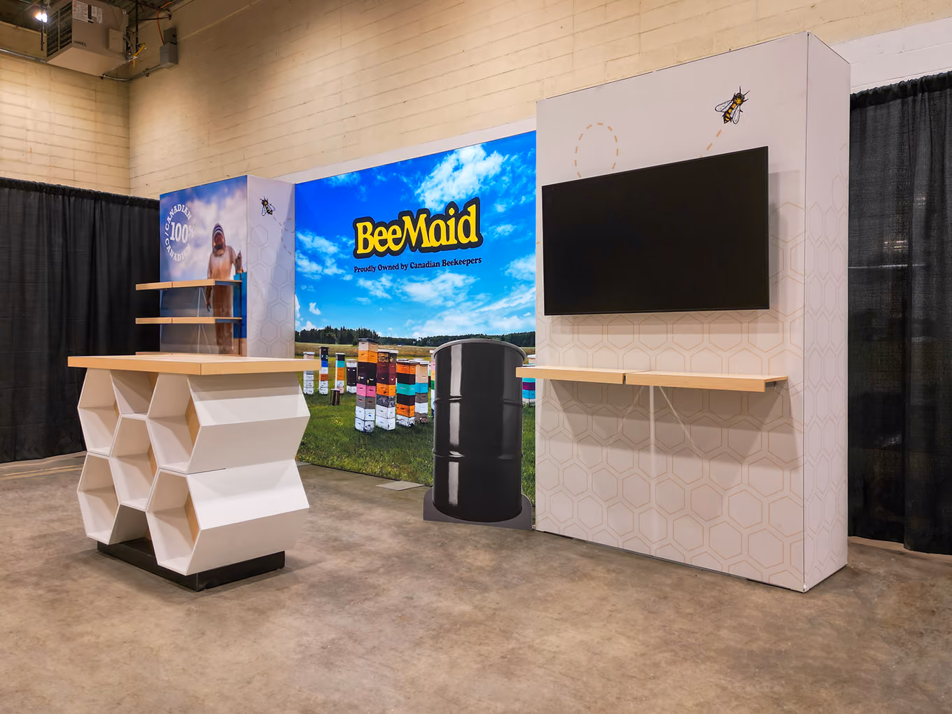 Angled view of BeeMaid Honey trade show display featuring custom honeycomb counter, honey barrel prop, and large-format "Proudly Owned by Canadian Beekeepers" brand mural.
