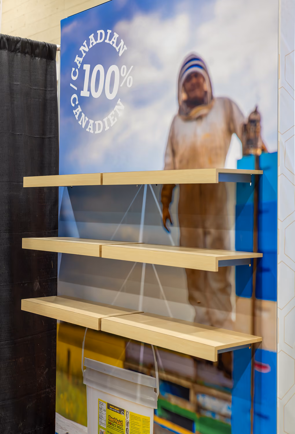Close-up of BeeMaid Honey trade show product shelving unit with bilingual "100% Canadian / Canadien" beekeeper graphic backdrop and maple floating shelves.