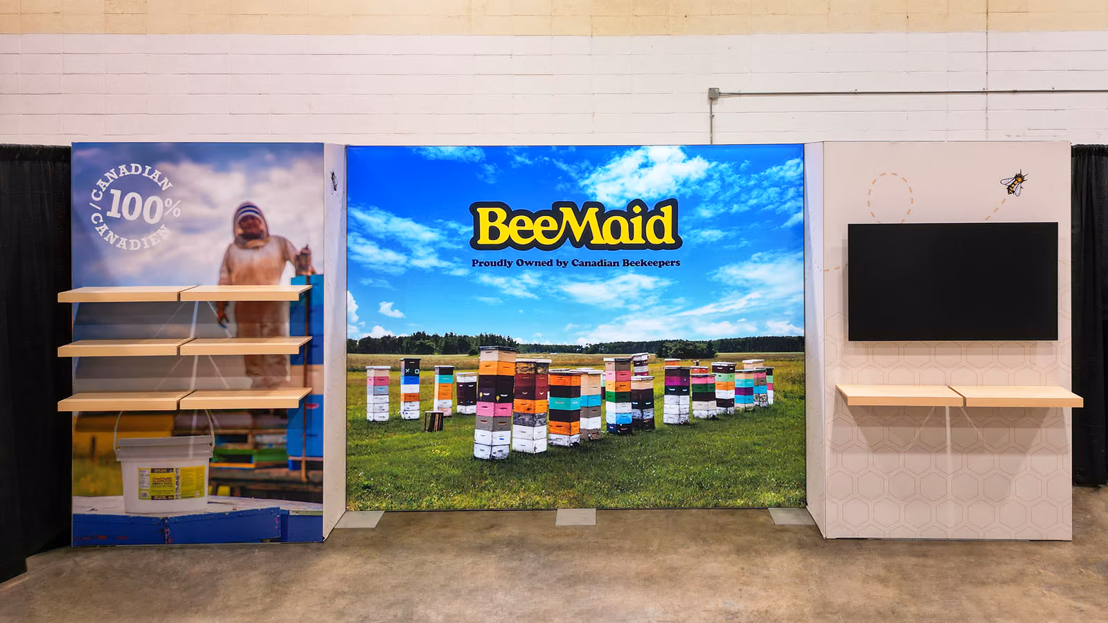 Full front view of BeeMaid Honey 10x20 trade show booth — backlit beehive field mural, beekeeper product shelf panel, hexagon-patterned monitor wall, and branded signage.