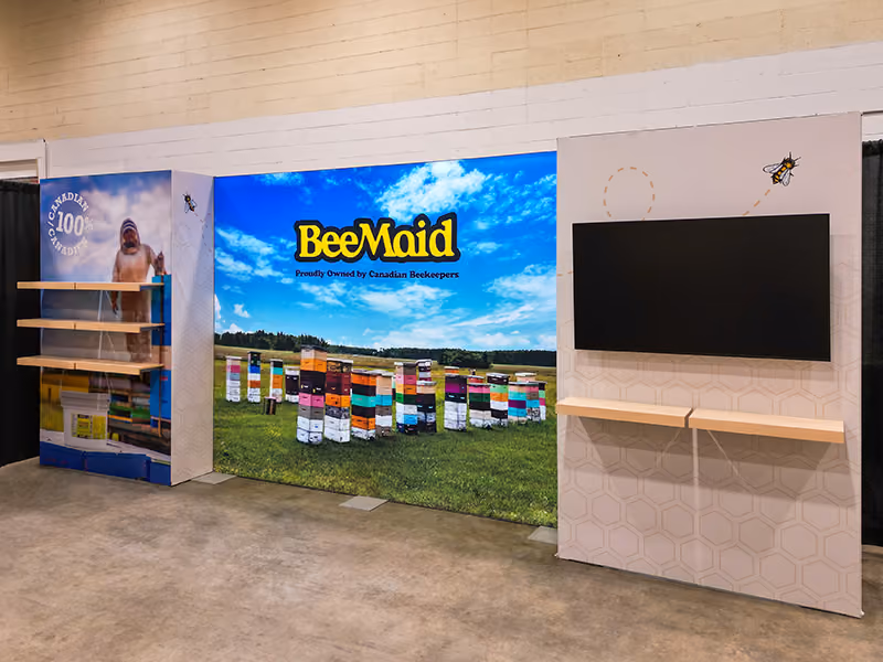 BeeMaid Honey 10x20 trade show booth without props — showing full backlit beehive field mural, beekeeper shelving panel, monitor wall, and honeycomb graphic panels.