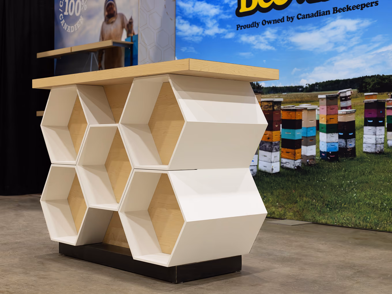 Custom honeycomb-shaped product display counter with maple top and white hexagonal shelving — BeeMaid Honey 10x20 trade show booth, Canadian honey brand.