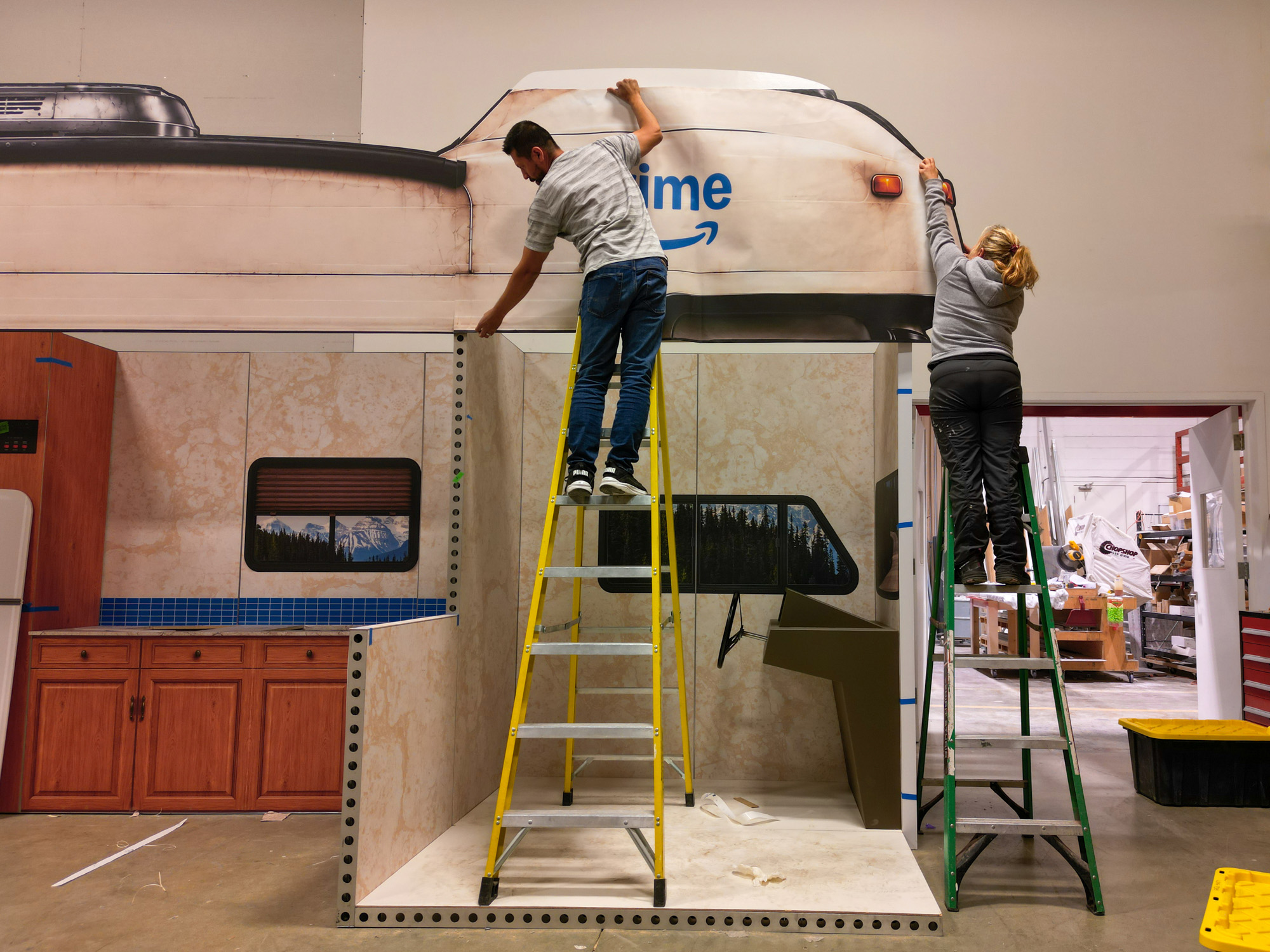 Two installers apply vinyl graphics to the roofline of the custom RV façade during on-site build.
