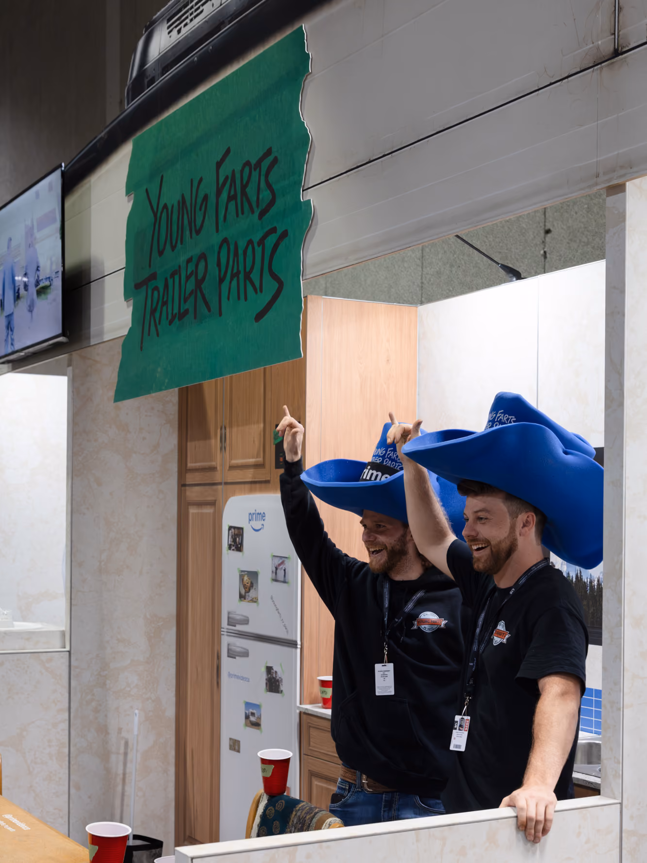 Cast members in blue foam hats point up at the Young Farts Trailer Parts sign inside the custom RV activation.