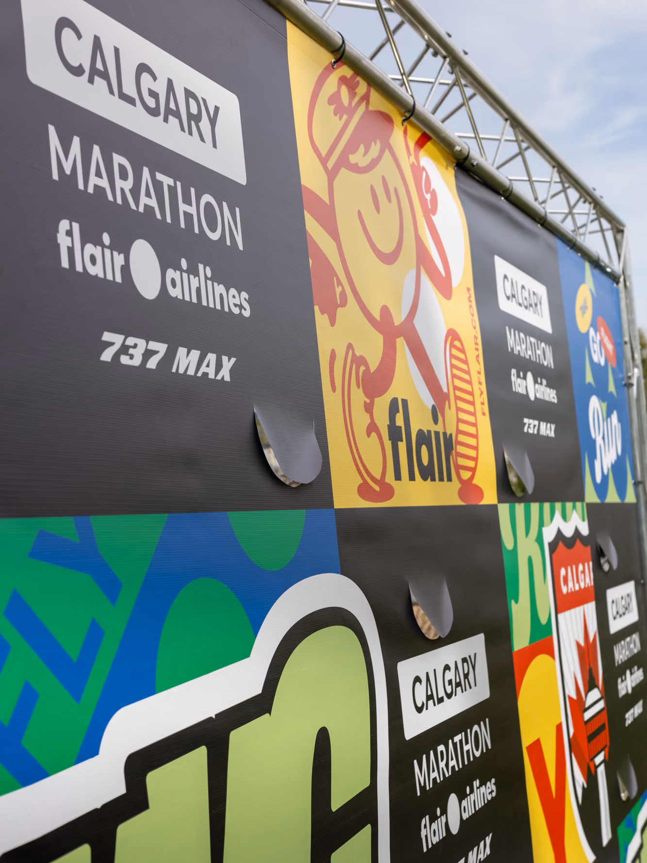 Angled close-up of printed vinyl graphics with wind slits and truss hooks, featuring Calgary Marathon and Flair Airlines branding.