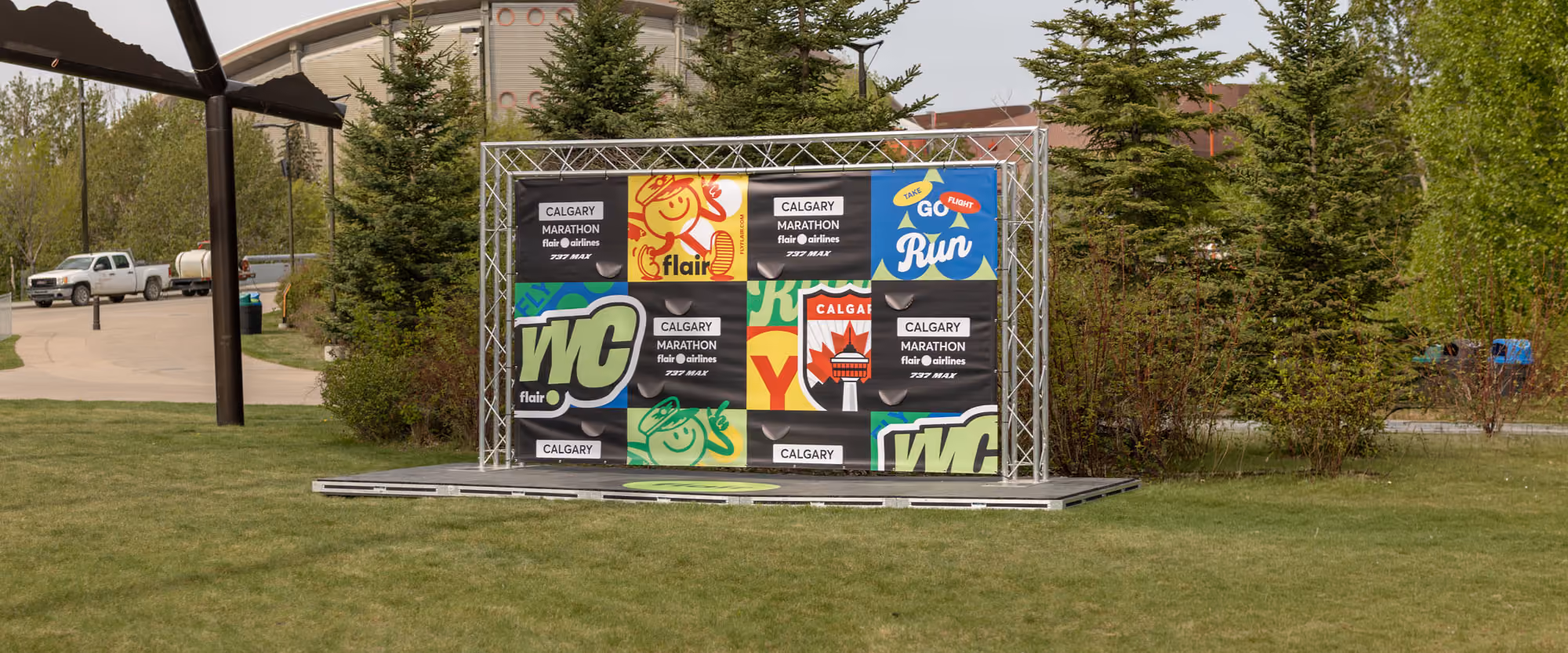 Flair Airlines and Calgary Marathon photo backdrop with colourful vinyl graphics on a truss and raised stage, set among spruce trees.