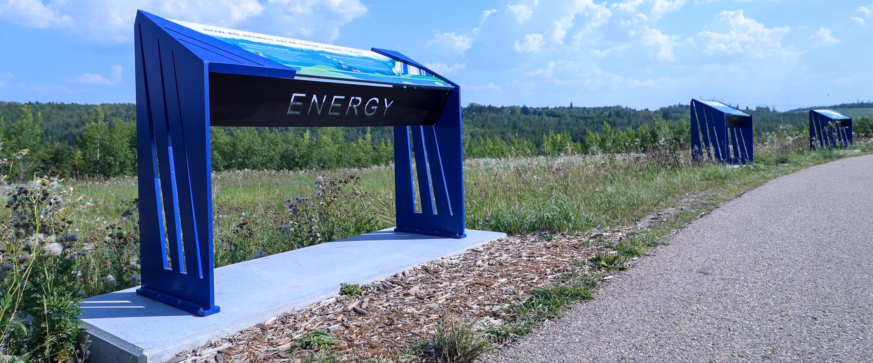Outdoor "Energy" interpretive panel along a gravel pathway, with a second sign visible in the distance.