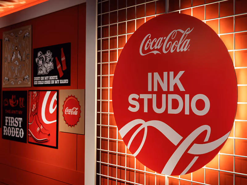 Backlit Coca-Cola Ink Studio circular sign mounted on a custom gridwall inside the branded seacan activation.
