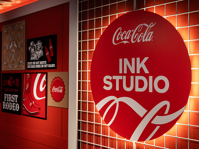 Backlit Coca-Cola Ink Studio circular sign mounted on a custom gridwall inside the branded seacan activation.