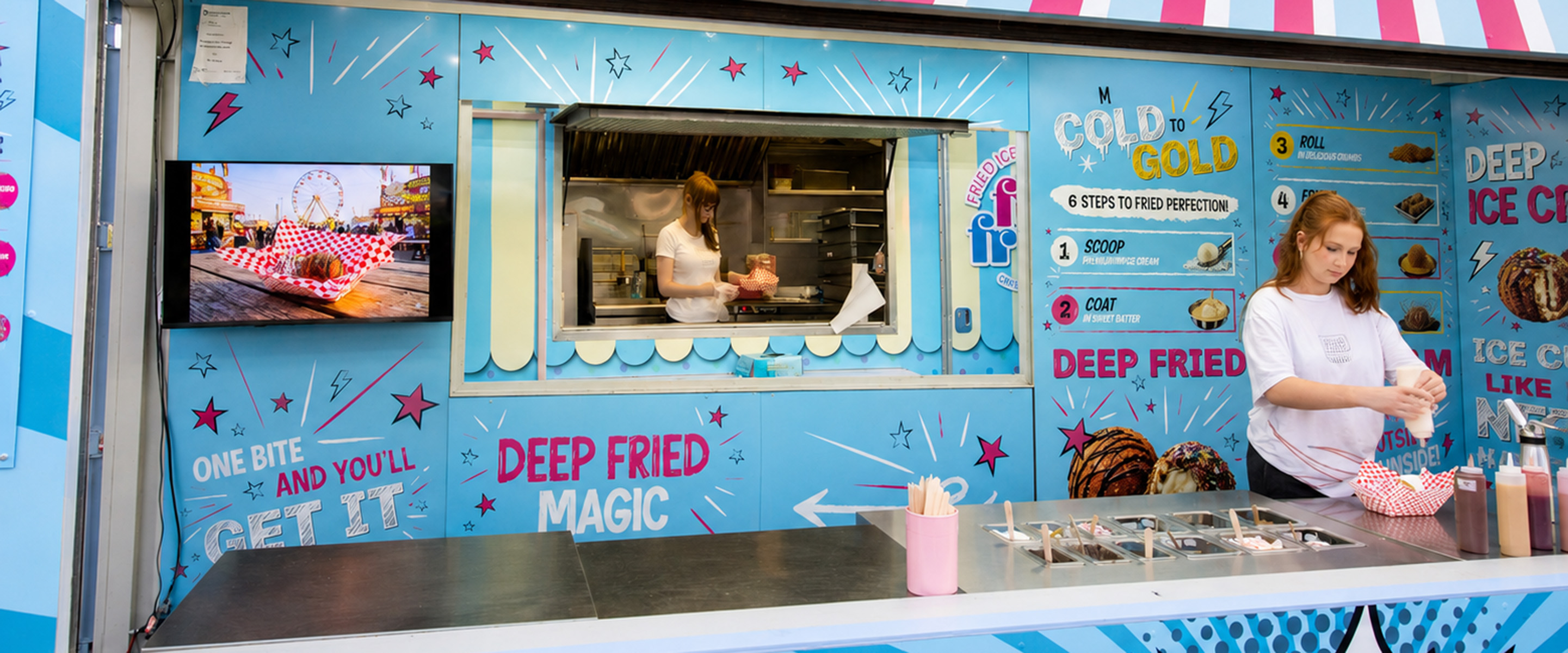 Inside the Frio Frio deep fried ice cream stand, where staff prepare orders at a topping station. The walls are covered in bright blue graphics, including a "Cold to Gold" step-by-step menu.
