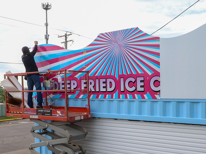 An Exhibit Studio installer on a scissor lift applies the "Deep Fried Ice Cream" graphic to the custom upper structure.