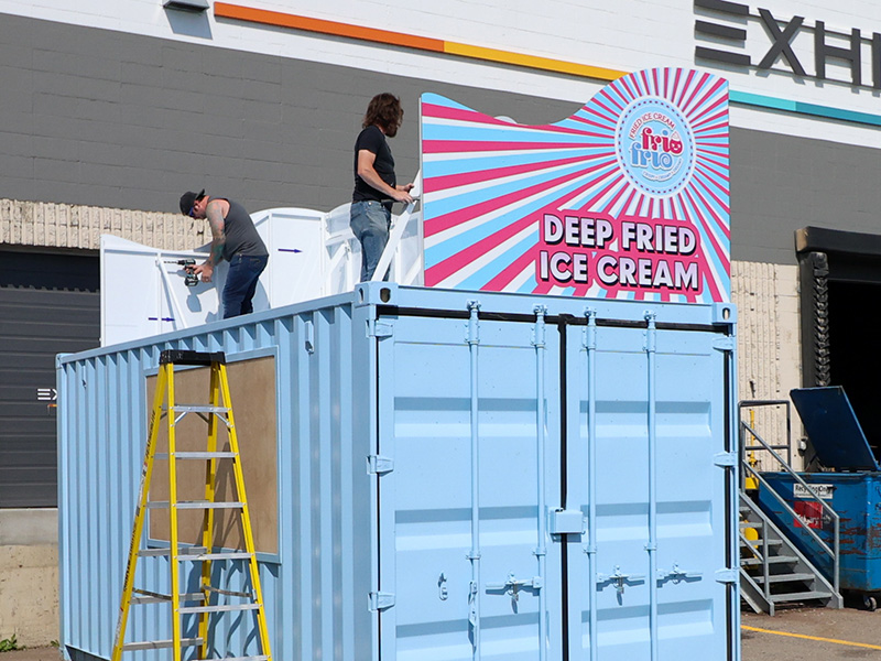 Two Exhibit Studio team members assemble the custom aluminum upper structure on the roof of a shipping container outside our shop.