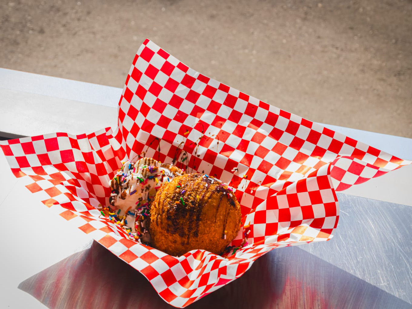 A scoop of deep fried ice cream topped with chocolate drizzle and rainbow sprinkles, served on red and white checkered paper.
