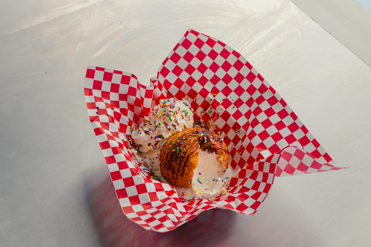 A scoop of deep fried ice cream with a bite taken out of it, topped with chocolate drizzle and rainbow sprinkles, served on red and white checkered paper.