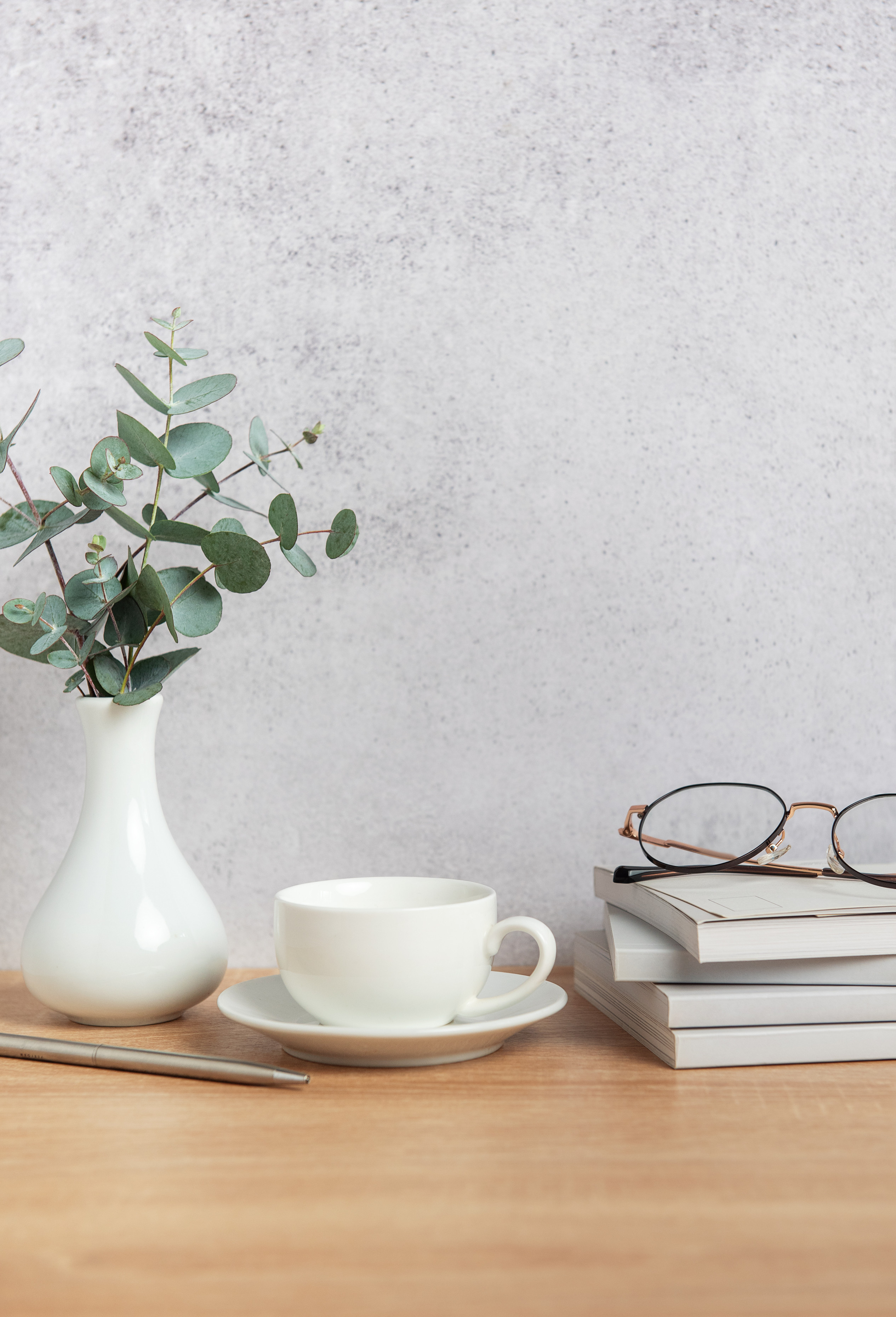 Minimalist desk with a cup of coffee, books, glasses, and a vase of greenery against a neutral wall.