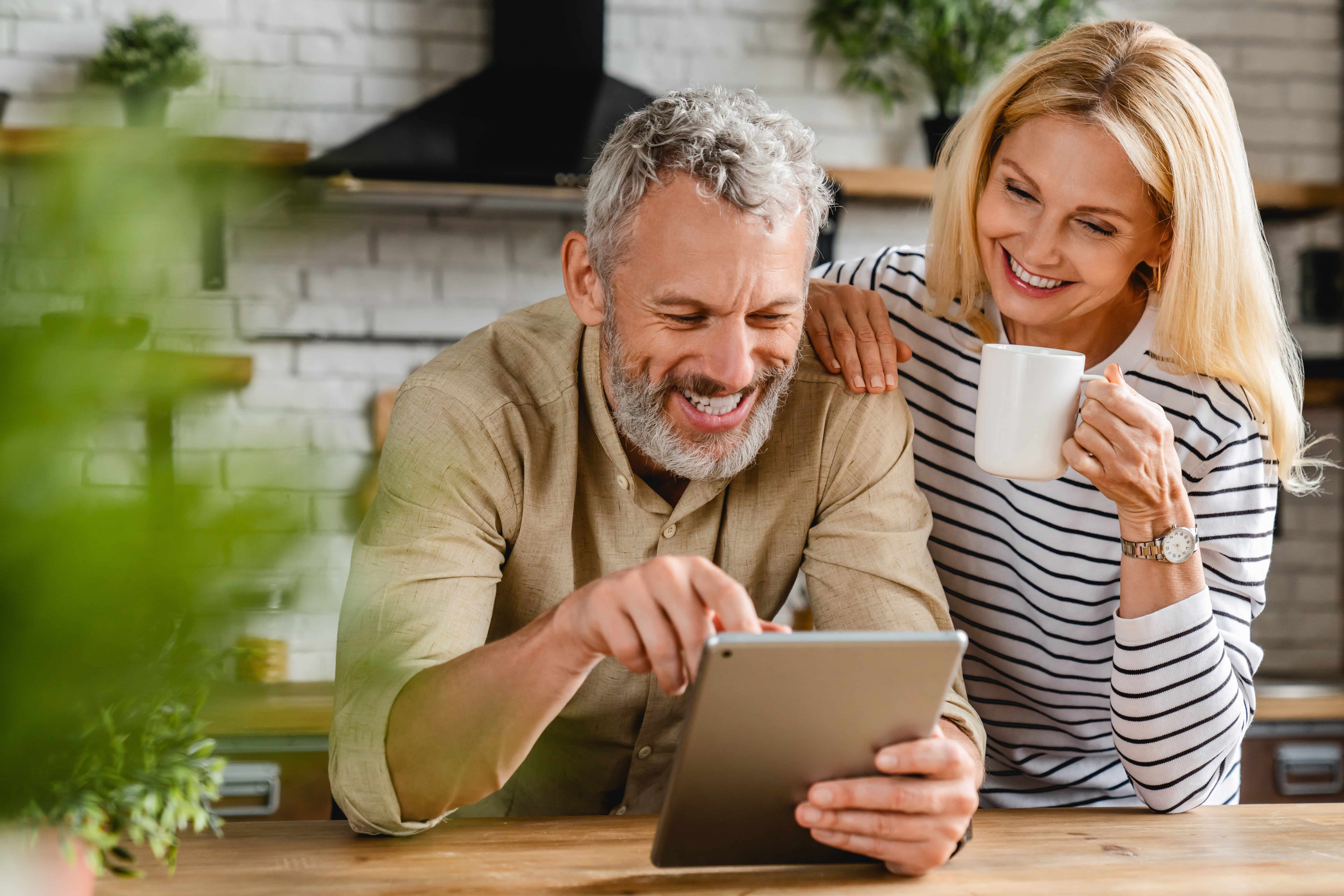 Smiling middle-aged man using a tablet while a woman in a striped shirt holds a white mug and looks on happily.