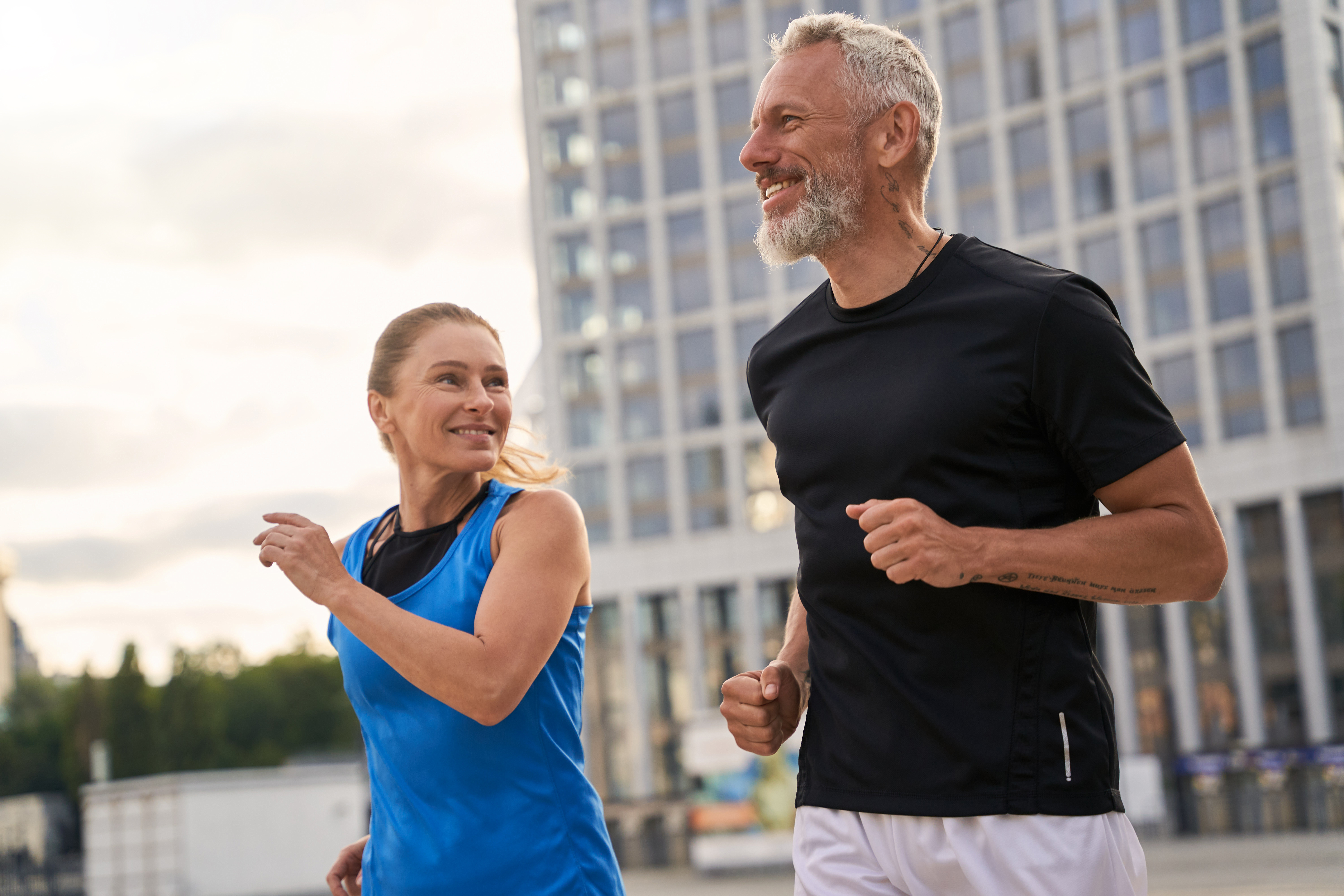Smiling man with gray beard in black shirt and woman in blue tank top jogging outdoors in an urban area.