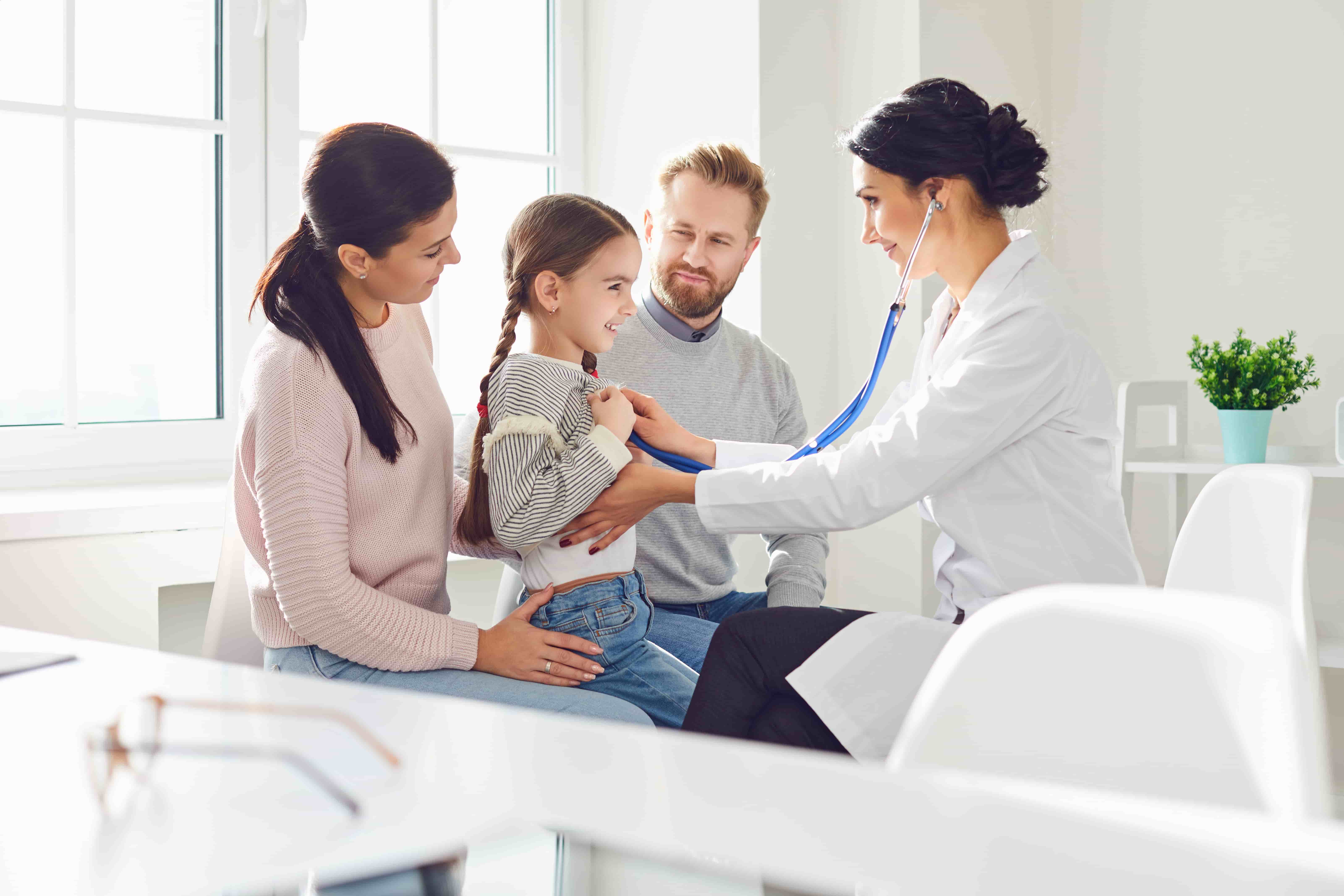 Female doctor using stethoscope to listen to a smiling young girl’s chest while her parents watch in a bright medical office.