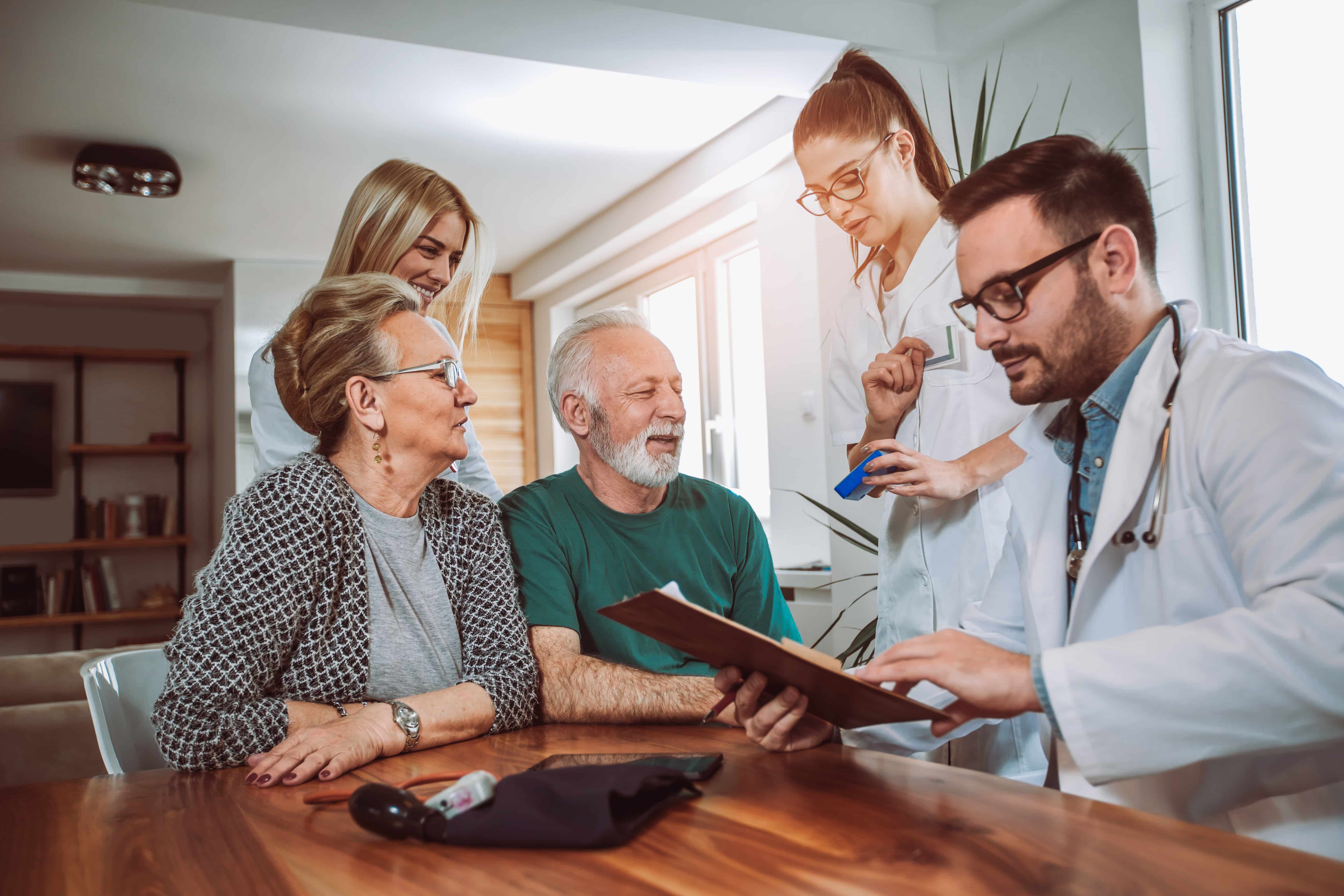 Two elderly adults seated at a table consulting with two doctors who are reviewing documents and medication in a bright room.