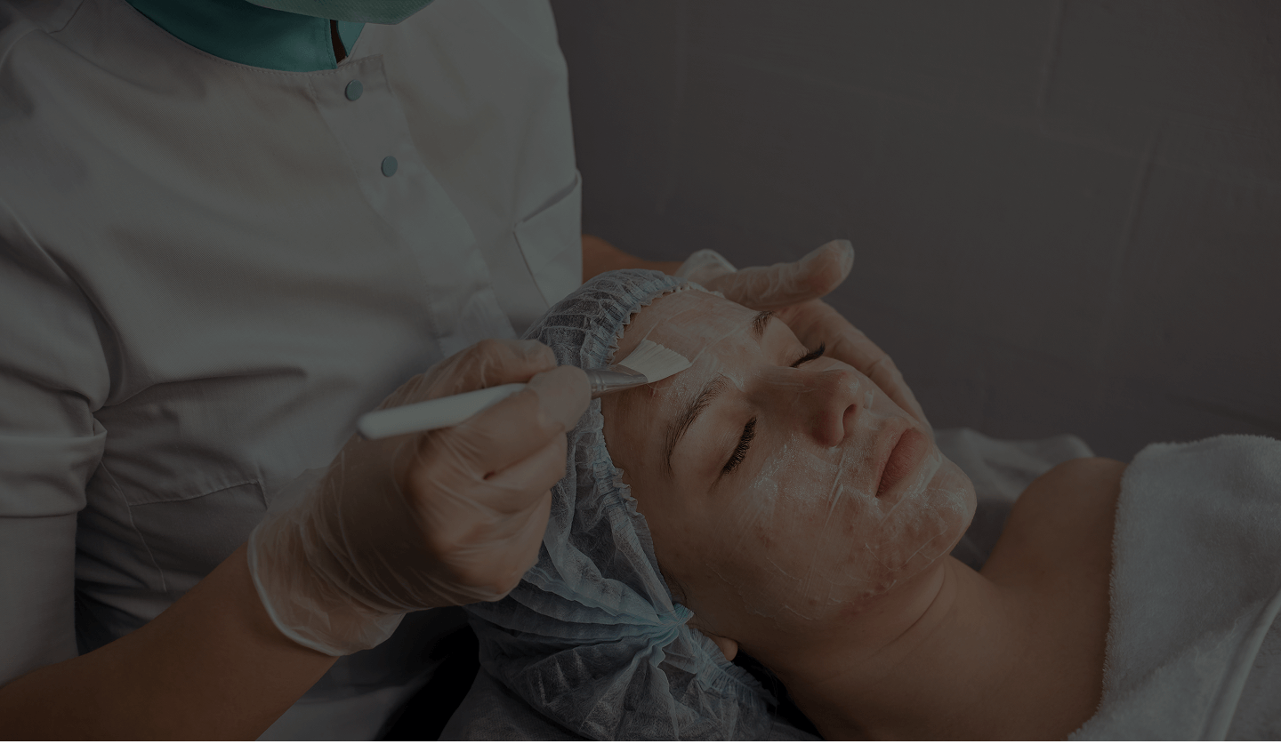 Person with protective gloves applying a facial mask on a woman’s face who is lying down with eyes closed and wearing a hair cap.