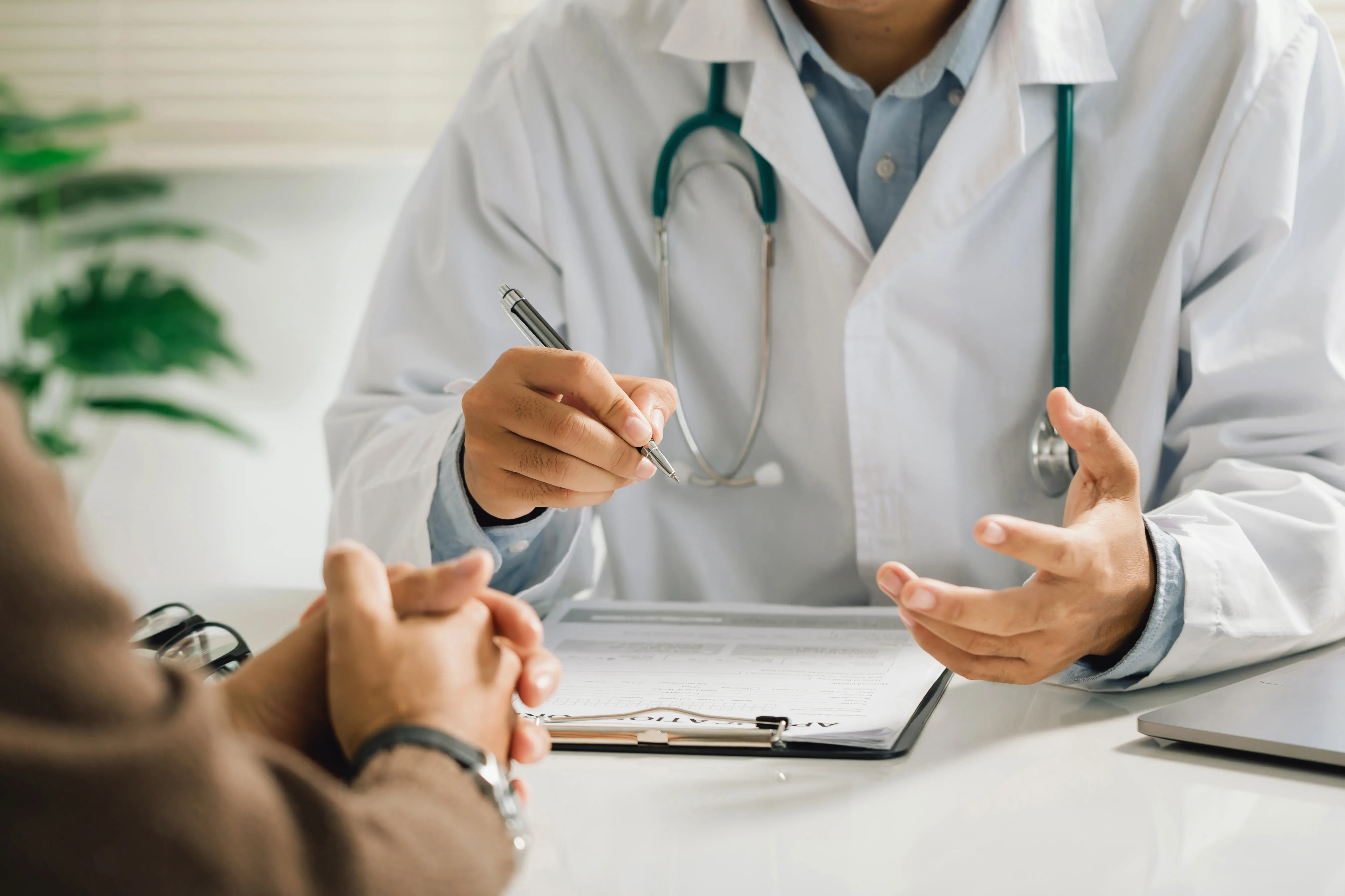 Doctor in a white coat with a stethoscope explaining medical information to a patient at a desk with a clipboard and documents.