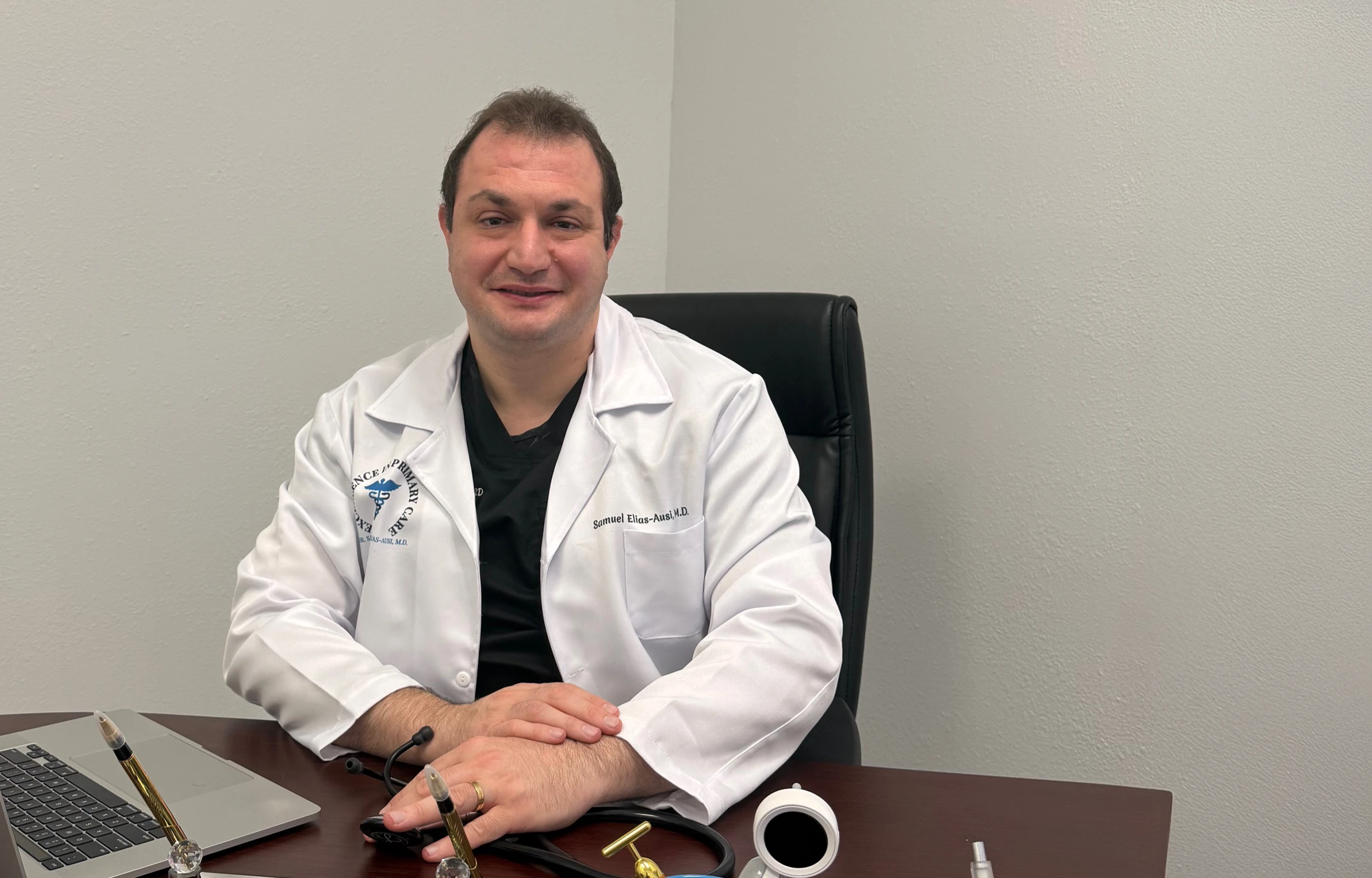 Smiling male doctor wearing a white coat sitting at a desk with a laptop and medical equipment.