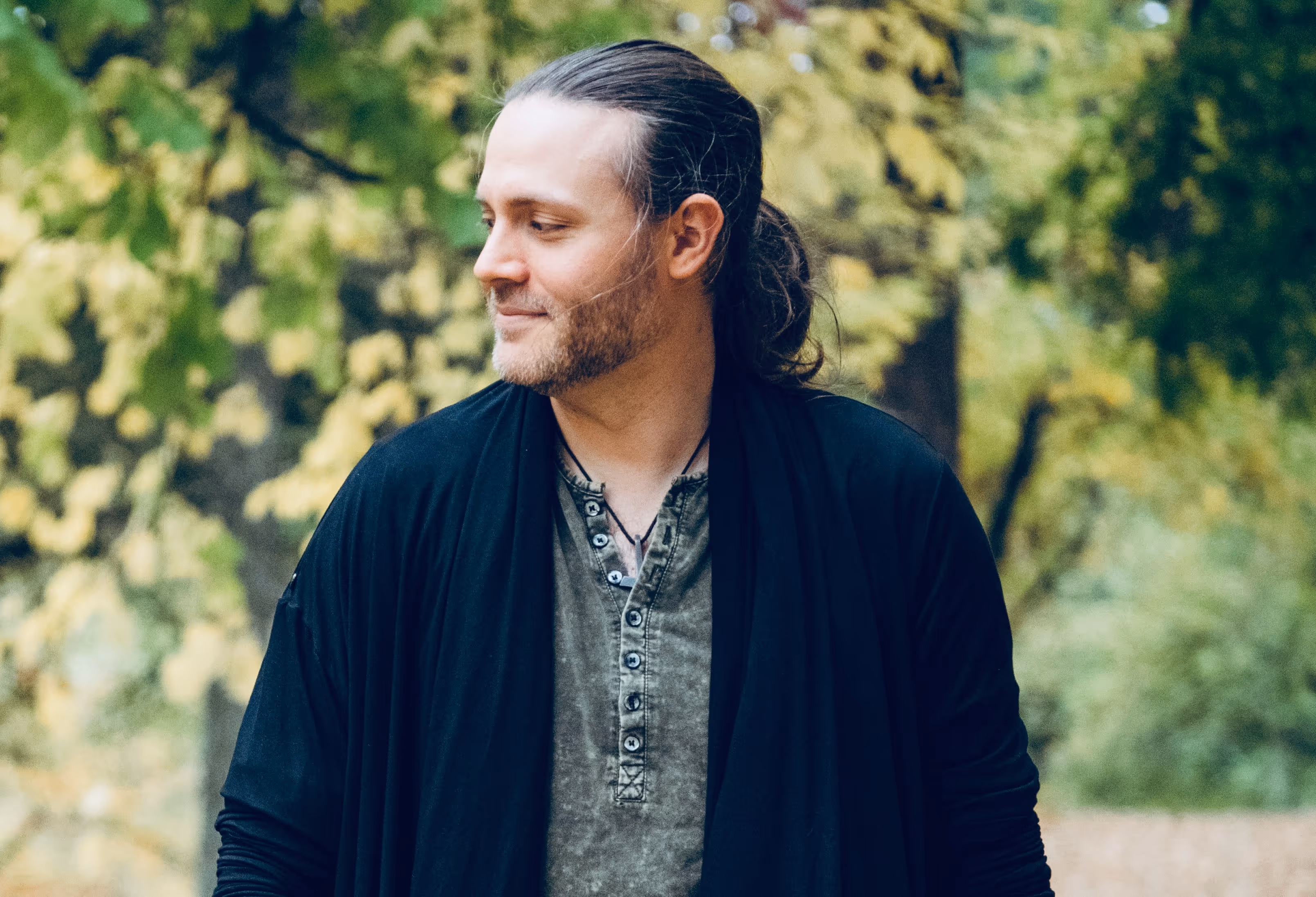 A man stands outdoors surrounded by soft green and yellow leaves, looking to the side with a calm, reflective expression. His posture and setting convey warmth, mindfulness, and grounded presence in nature.
