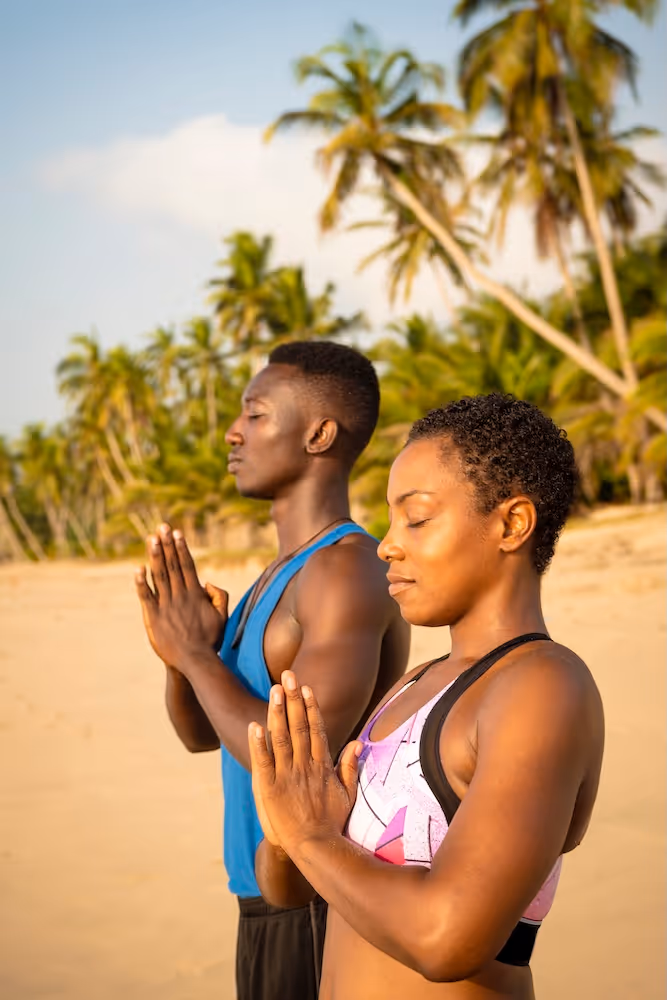 Two people stand in prayer on a golden beach, eyes closed, palms together, surrounded by palm trees and ocean breeze—embodying calm, gratitude, and grounded focus.

