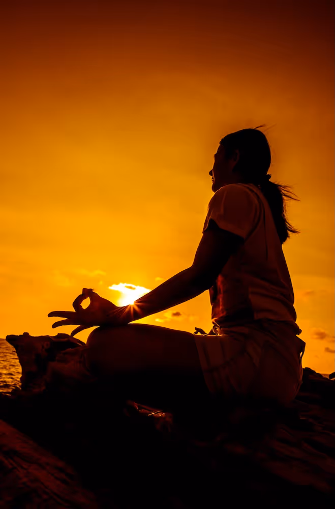 A person meditates at sunset, silhouetted against an amber sky, fingers forming a mudra—evoking stillness, breath, and the meeting of inner and outer light.
