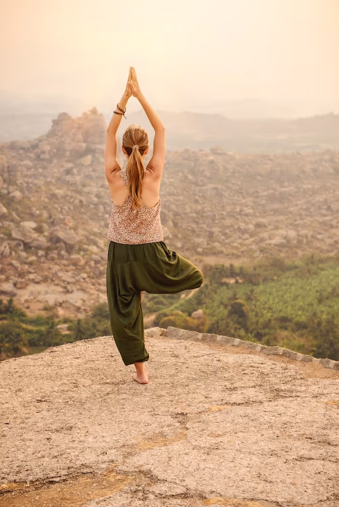 A woman practices yoga in tree pose at the edge of a rocky overlook, sunlight wrapping her form as the landscape stretches endlessly below—balance and openness in motion.
