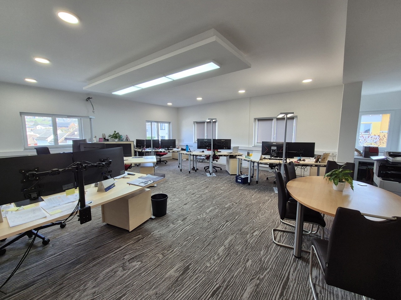 Helles Büro mit mehreren Schreibtischen, Computermonitoren, einem runden Tisch mit vier Stühlen und einem kleinen Pflanzentopf.