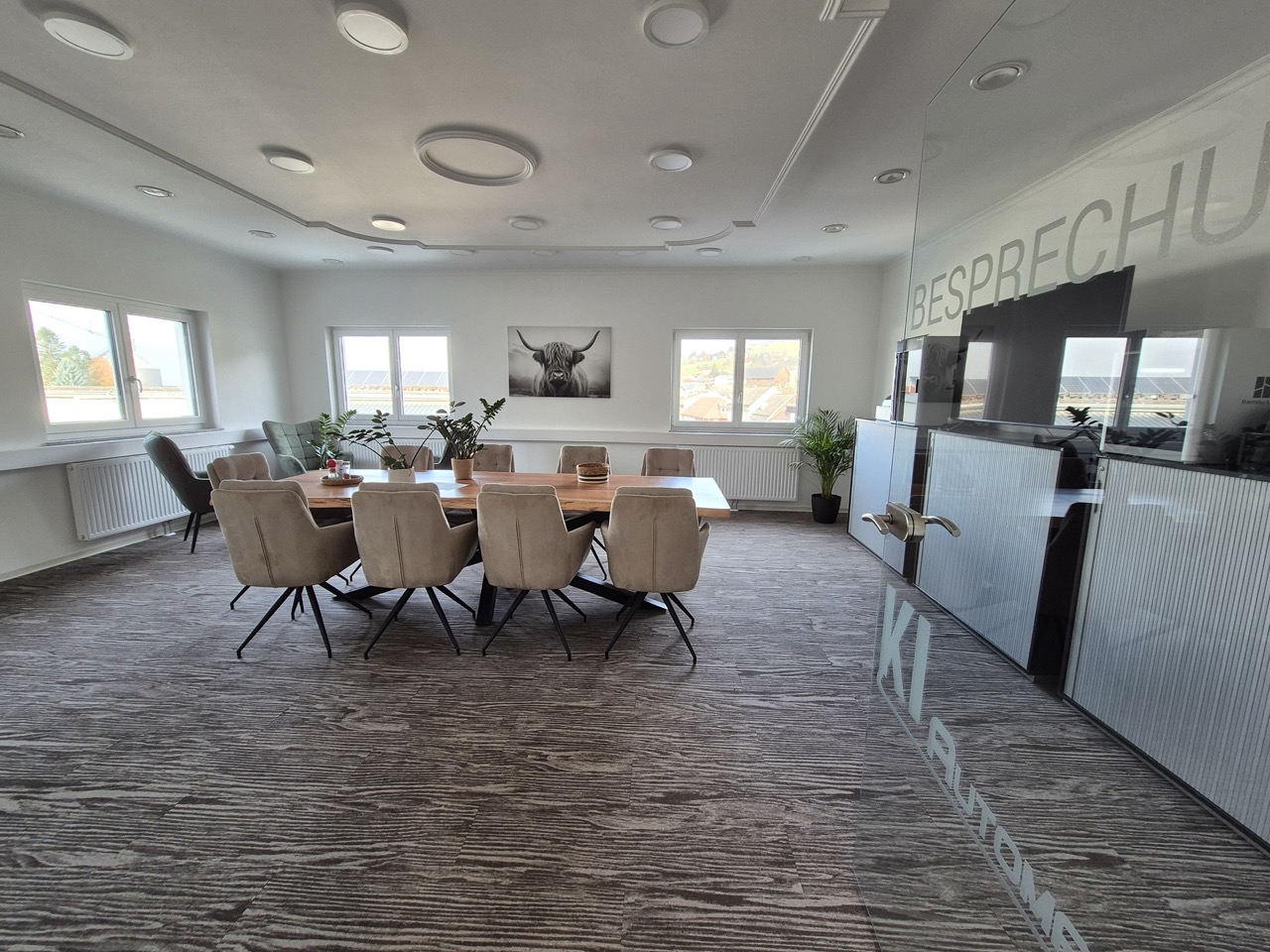 Moderner Besprechungsraum mit großem Holztisch, beigen Stühlen, mehreren Pflanzen und drei Fenstern mit Blick nach draußen.