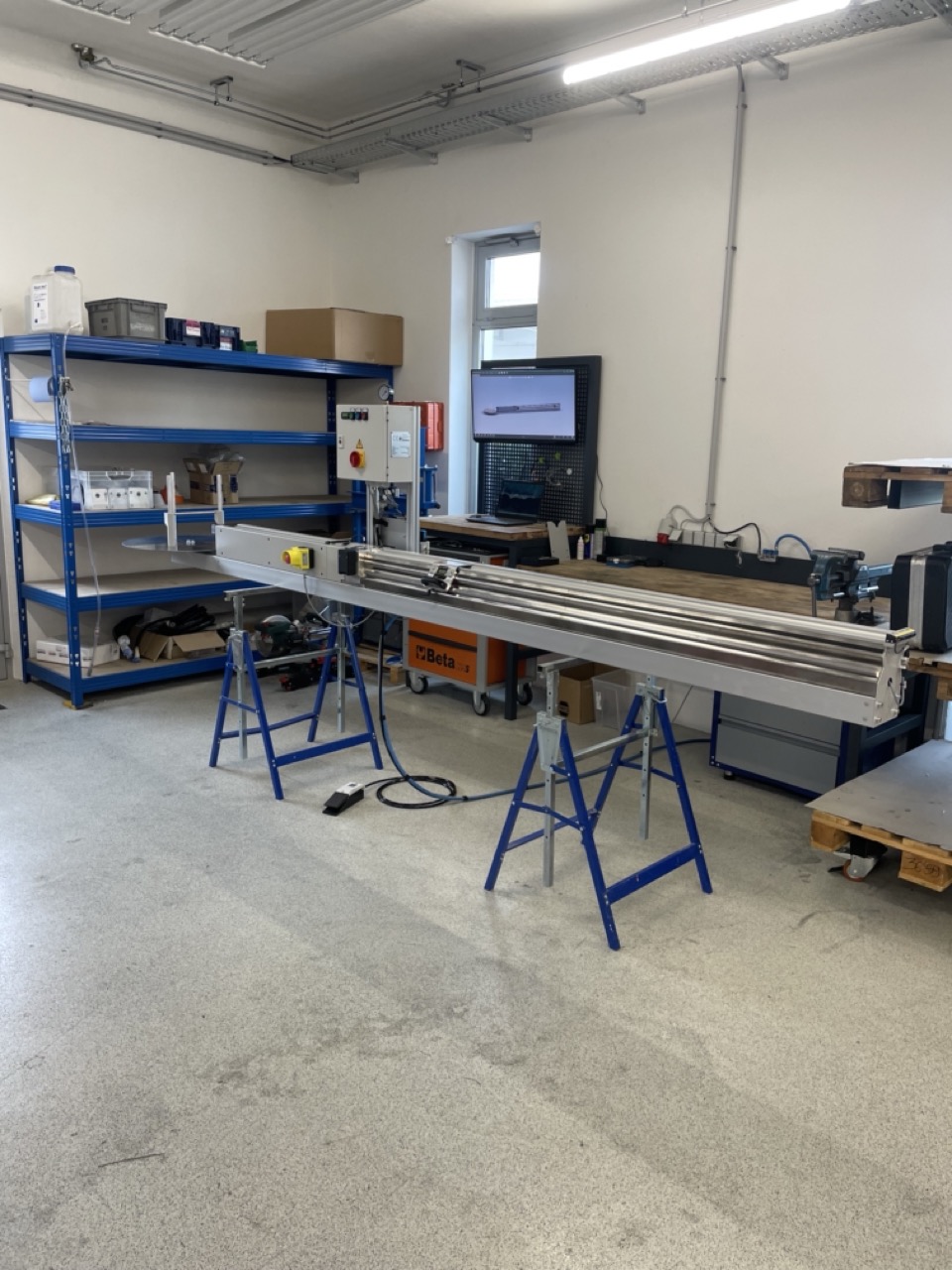 Werkstatt mit einer langen Metallmaschine auf zwei blauen Böcken, Regalen mit Werkzeugen und einem Monitor an der Wand.