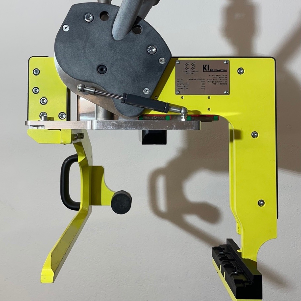 Gelbes KI Automation-Haltevorrichtung mit Metallbeschlägen und schwarzem Griff an einer weißen Wand.