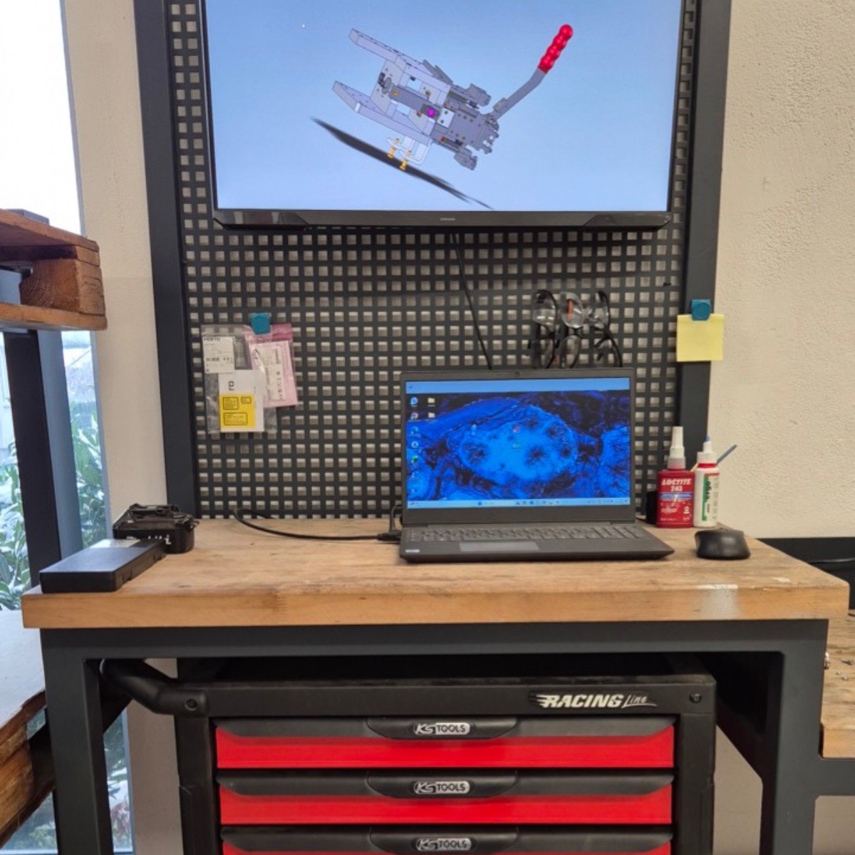 Werkbank mit Laptop, Computermaus, Klebstoffflaschen und einer an der Wand montierten Anzeige mit technischer Zeichnung.