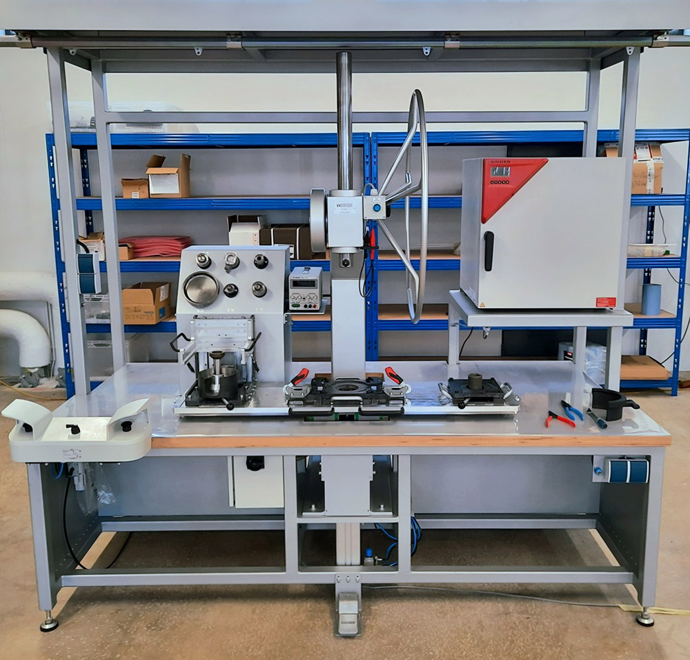 Industrieller Labortisch mit Prüfmaschine, Ofen und Werkzeugen vor Regalen im Lagerraum.