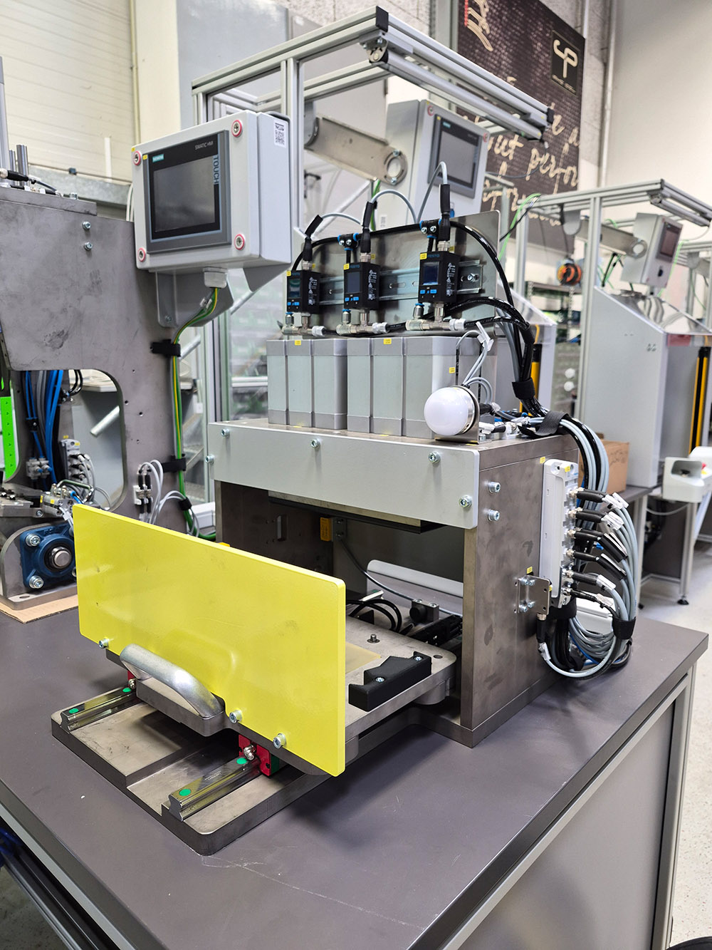 Industrielle Maschine mit Steuergerät und gelbem Sicherheitsschutz auf grauem Tisch in Werkstatt.