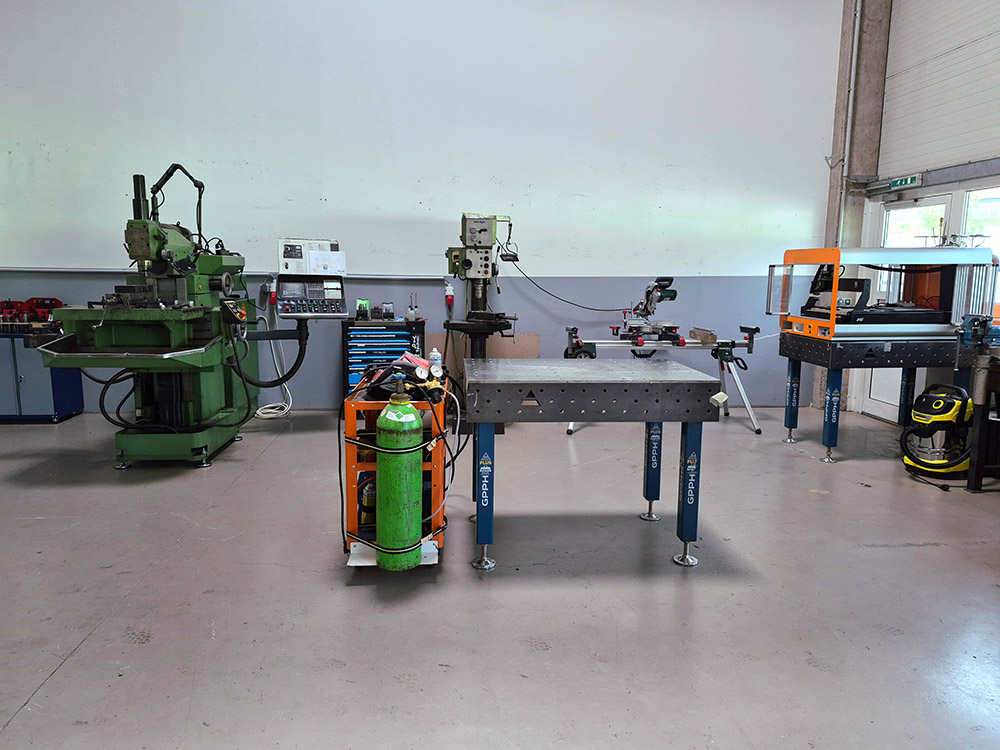 Werkstatt mit Maschinen, Gasflasche und Werkbank auf grauem Boden vor weißer Wand.