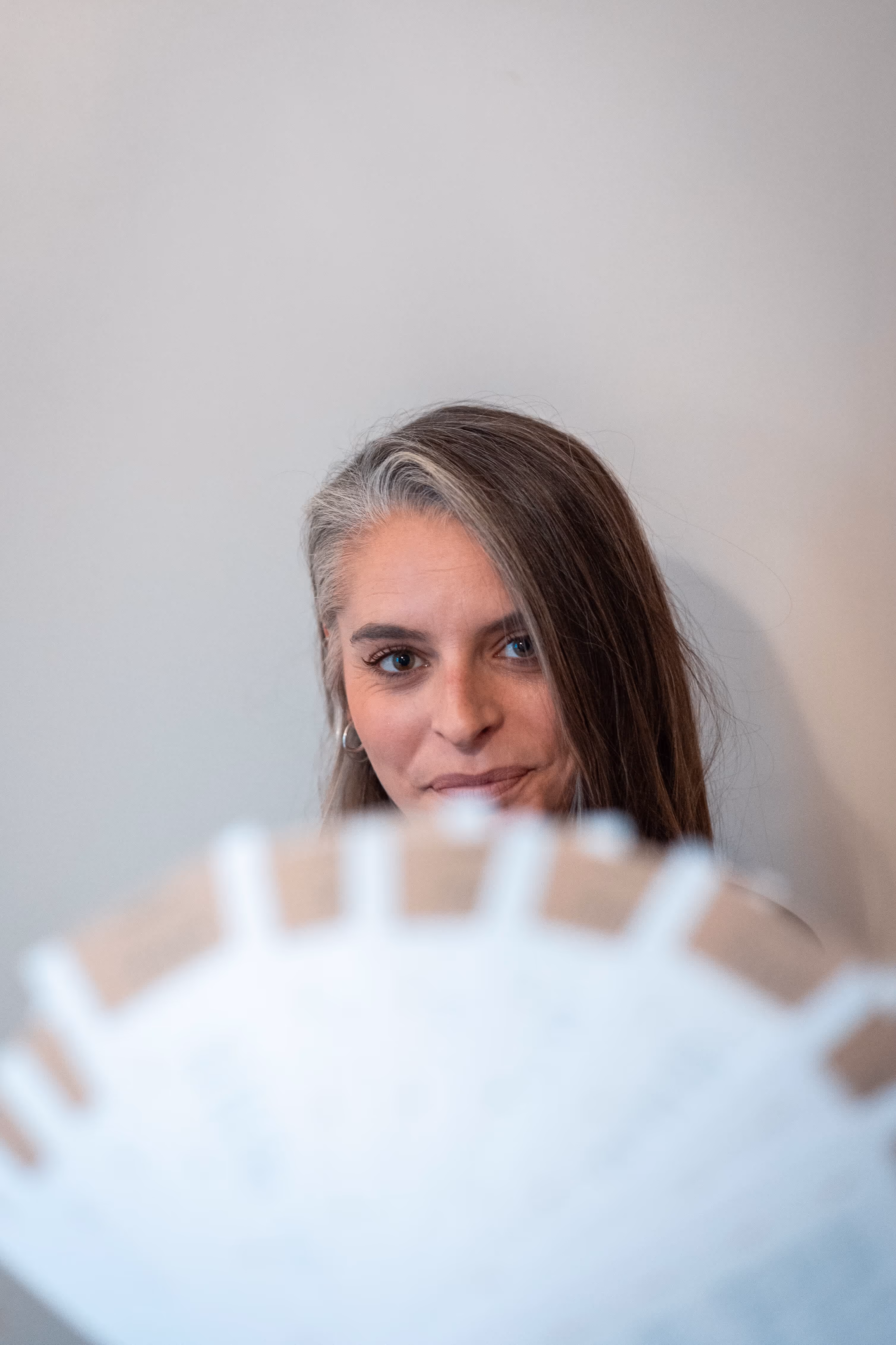 Portrait en plan moyen d'une femme aux cheveux bruns et gris, regardant directement l'appareil photo avec un léger sourire. Une partie floue d'un objet en forme d'éventail ou de demi-cercle, de couleur beige et blanc, masque le bas de son visage au premier plan. L'arrière-plan est un mur blanc uni.