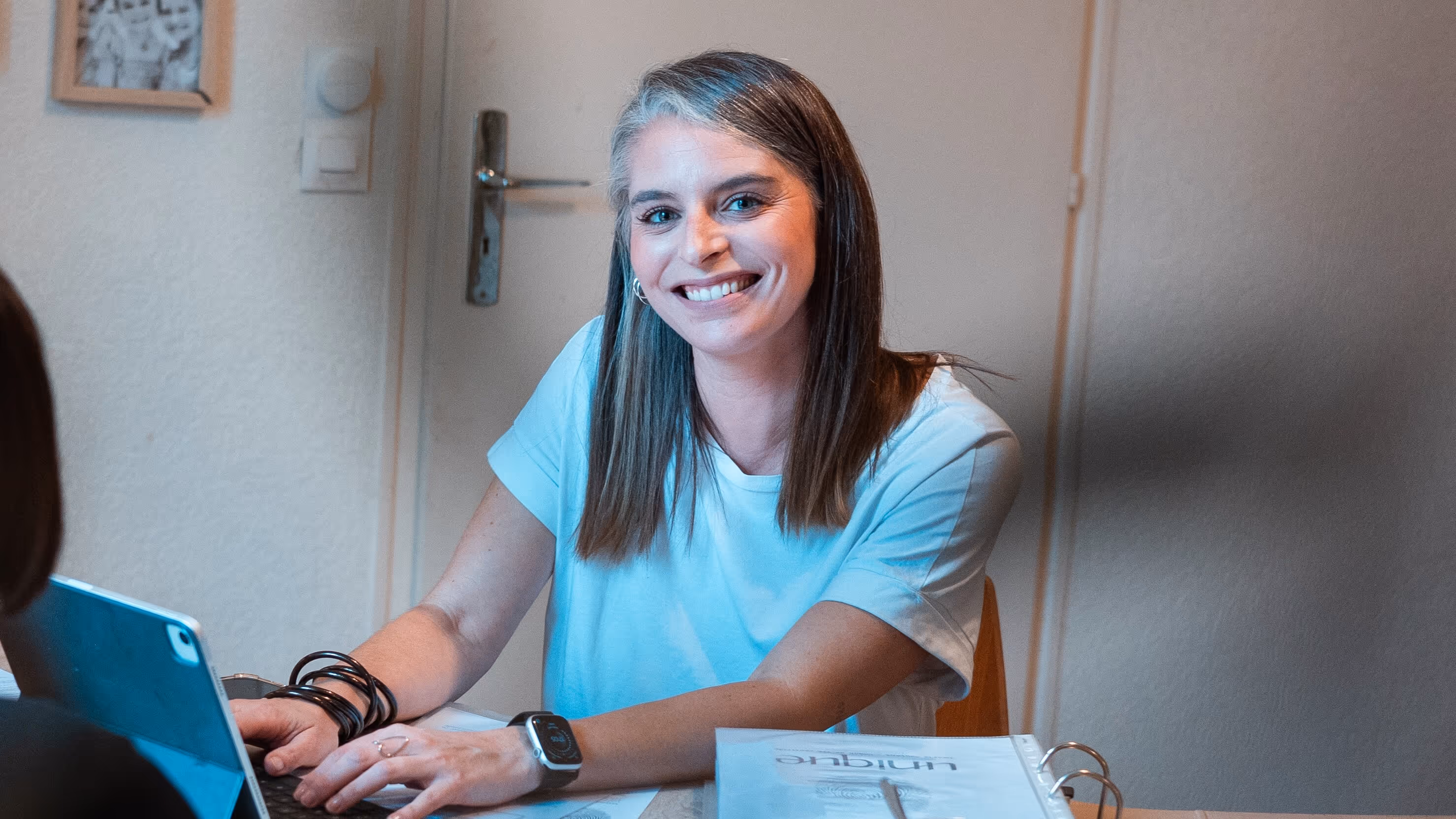 Deux femmes discutant à une table en bois dans une pièce décorée de photos encadrées, l’une souriant avec un ordinateur portable devant elle.