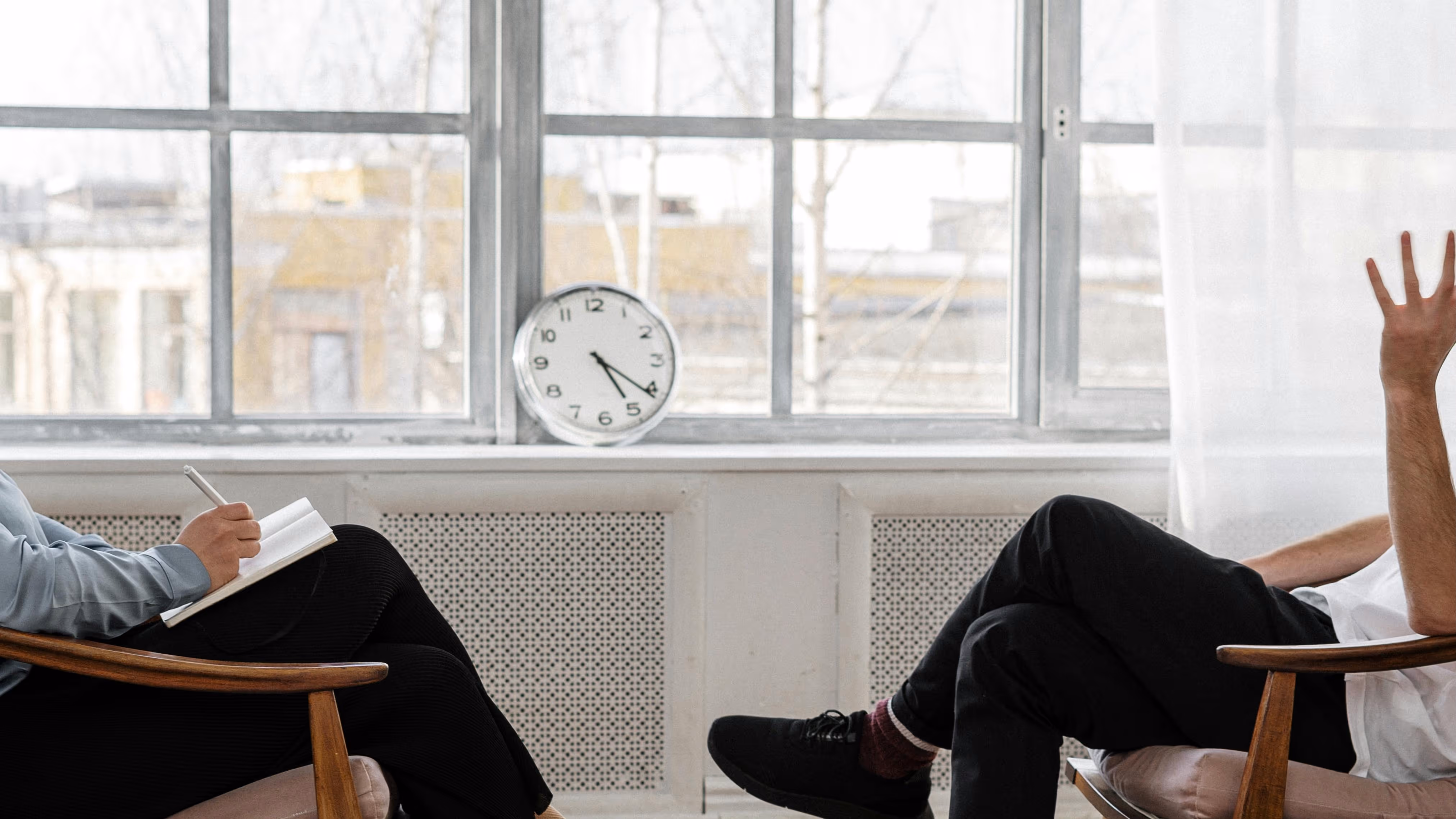 Plan moyen et horizontal de deux personnes assises l'une en face de l'autre dans des fauteuils en bois, se coupant au niveau du torse. La personne de gauche écrit dans un carnet. La personne de droite est vue de profil, levant un bras en l'air, la paume ou