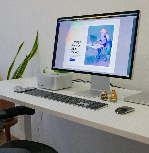 Minimalist desk setup with a monitor displaying a page titled 'Change the way art is valued,' keyboard, mouse, small figurines, and a plant in the background.