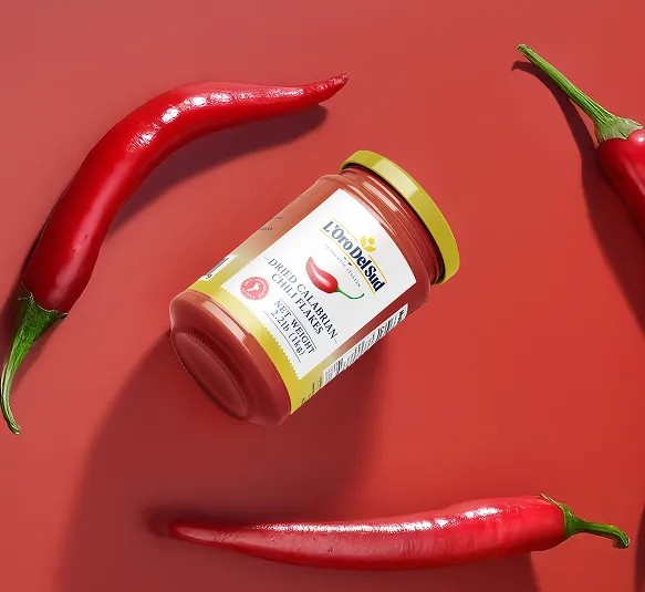 Jar of L'Oro Del Sud dried Calabrian chili paste surrounded by three whole red chili peppers on a red background.