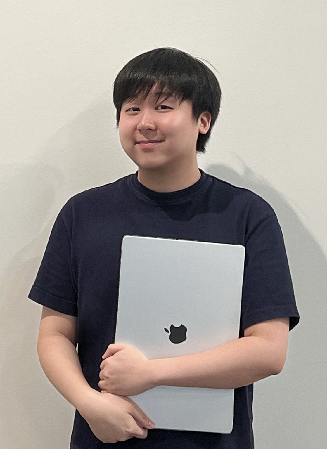 Young man with black hair wearing a dark t-shirt, smiling and holding a closed silver Apple laptop.