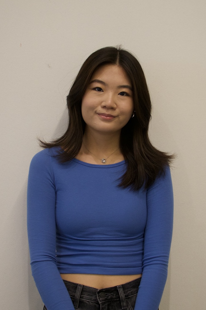 Young woman with long dark hair wearing a blue long-sleeve crop top and black jeans standing against a plain light-colored wall.