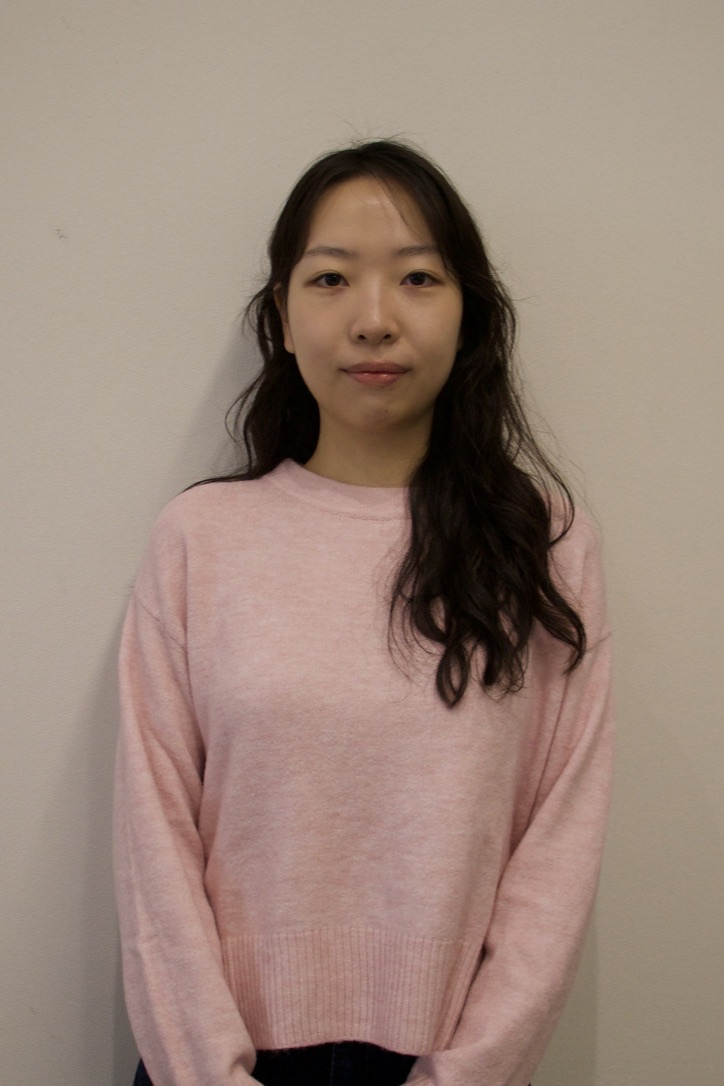Young woman with long black hair wearing round glasses, a purple sweatshirt, a necklace, and a bracelet, standing against a plain wall.